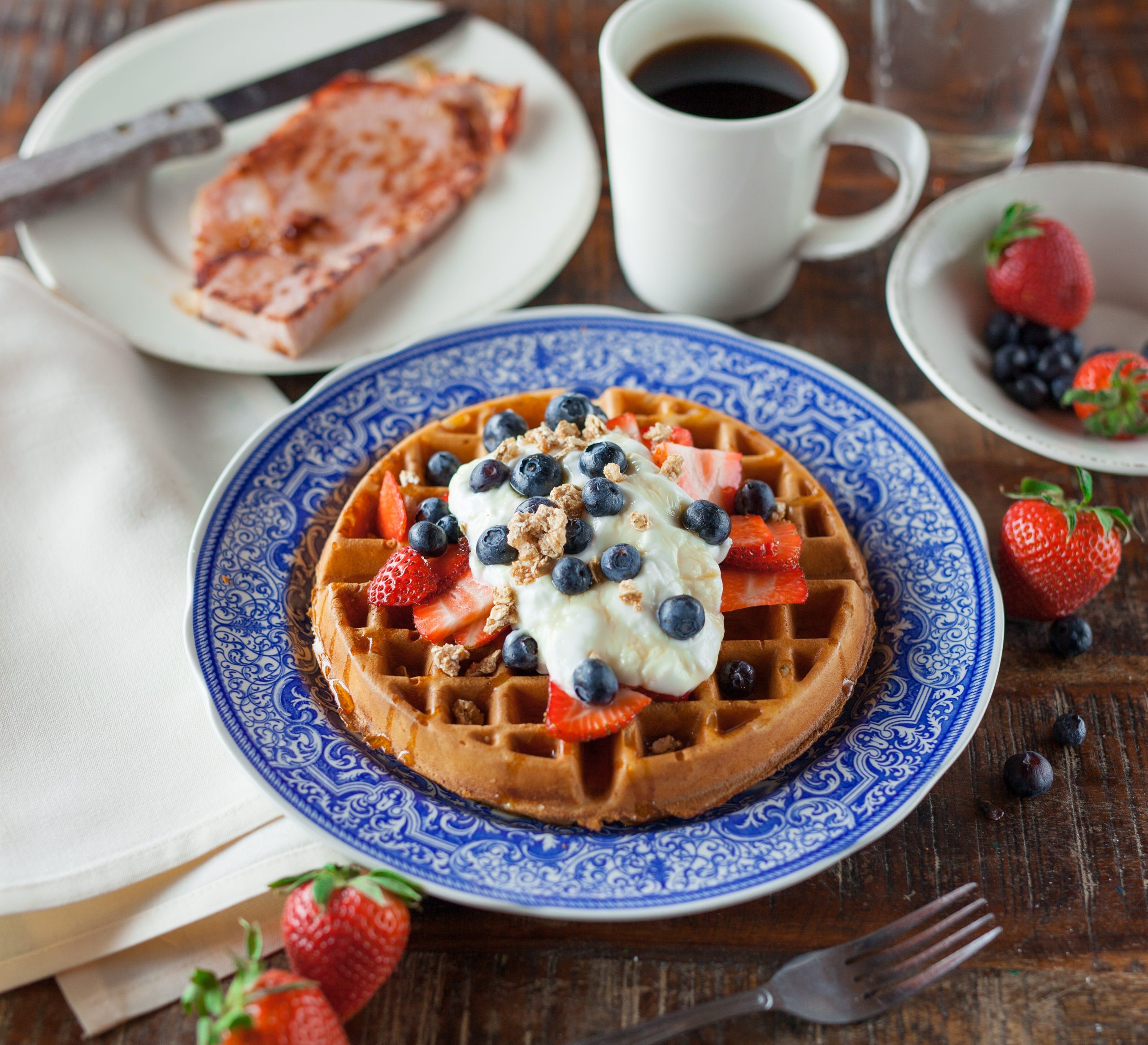 A plate of waffles surrounded by strawberries, ham, and coffee.